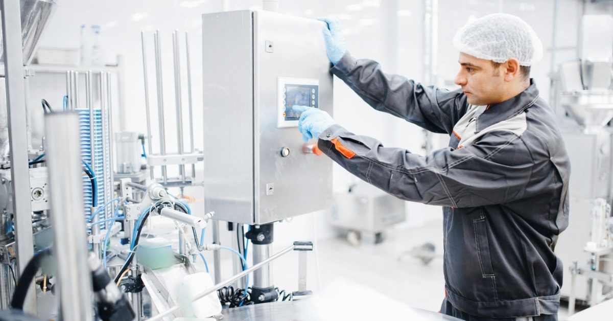 A person wearing a hairnet and gray uniform uses a control panel beside metal equipment in a bright factory.