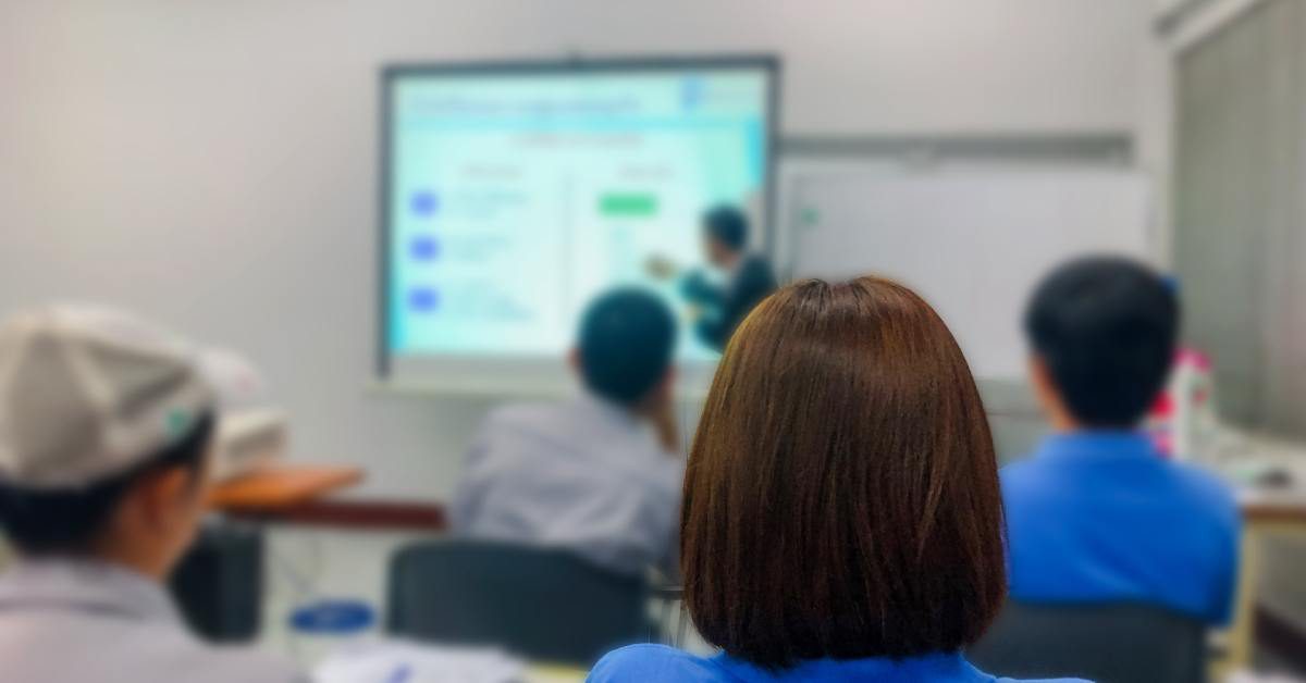 Workers sit at tables in a conference room and watch someone point at a presentation on a projector screen.
