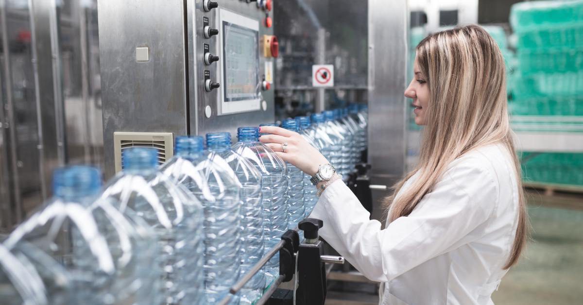 A female factory worker inspects a water bottle as it moves horizontally with others on a packaging machine.