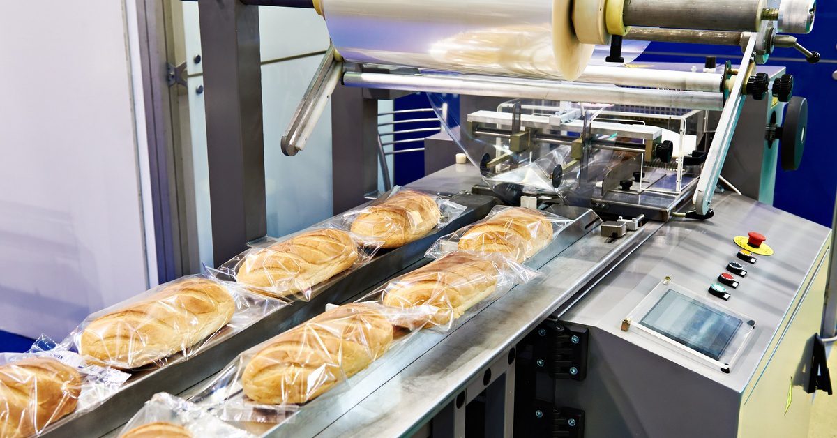 A horizontal packaging machine with a large, clear film roll processes multiple packages of bread through a conveyor.