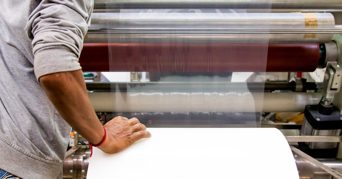 A person in a gray hoodie positions a strip of clear film through the rollers on a packaging machine.