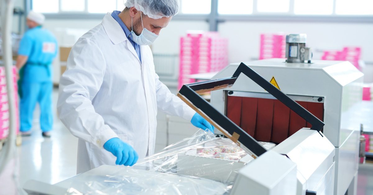 A factory worker wearing a hairnet and mask inspects the film on a box as it moves through a packaging machine.