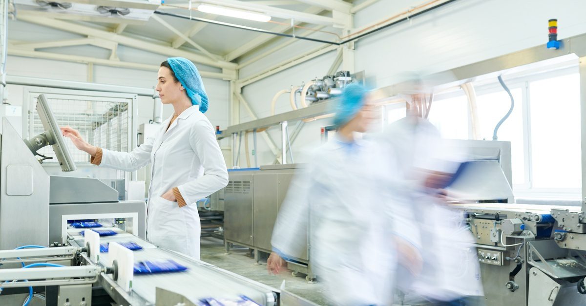 A woman in a white coat and a blue hairnet touches a screen above a packaging machine that processes products.