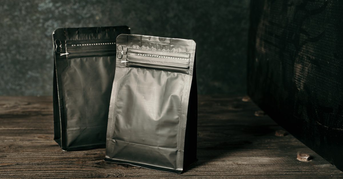 Two pouch packages, one black and the other dark gray, are placed on a wooden surface under soft lighting.