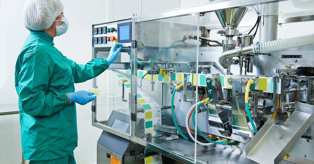 A worker wearing protective gloves and a facemask stands near a machine with transparent glass panels.