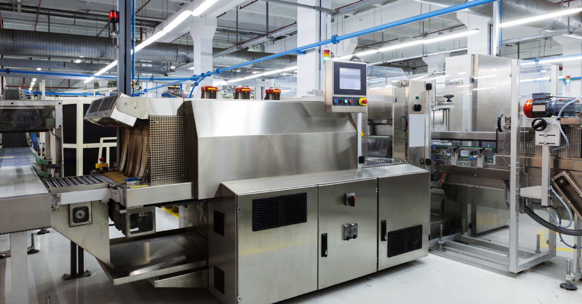 Stainless steel machines in a bright factory, with conveyors, control panels, and overhead ducts in a production line.