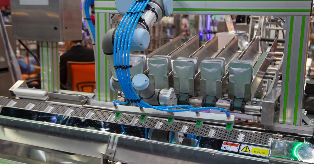 A robotic arm with blue cables operates above a conveyor carrying gray pouches in a well-lit industrial setting.