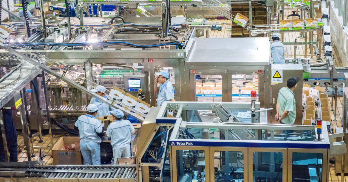Overhead view of a busy packaging factory with conveyor belts, machines, workers in blue uniforms, and boxed products.