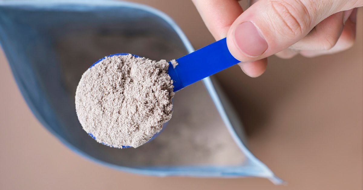 A hand holds a blue scoop of light-brown powder over an open, light-blue pouch, set on a plain beige surface.
