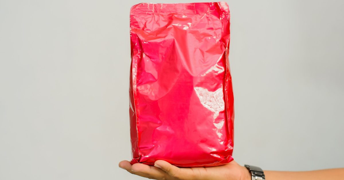 A hand with a wristwatch holds a shiny, crinkled red foil pouch upright against a plain gray background.
