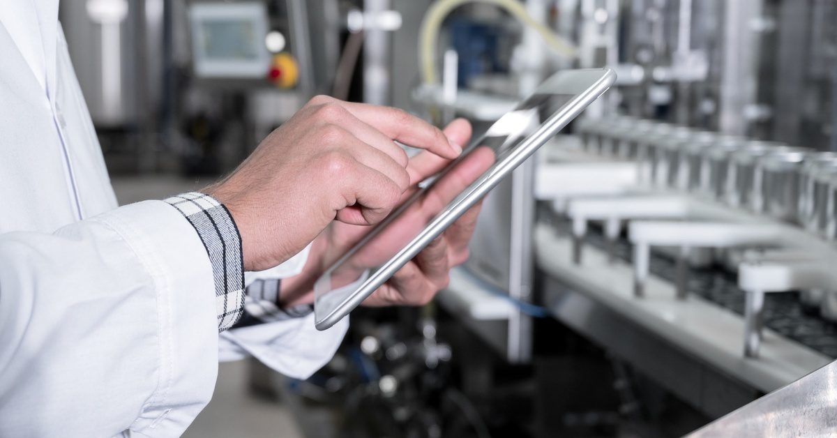 A person in a lab coat uses a tablet in a bright lab with blurred machinery and rows of small, clear containers.