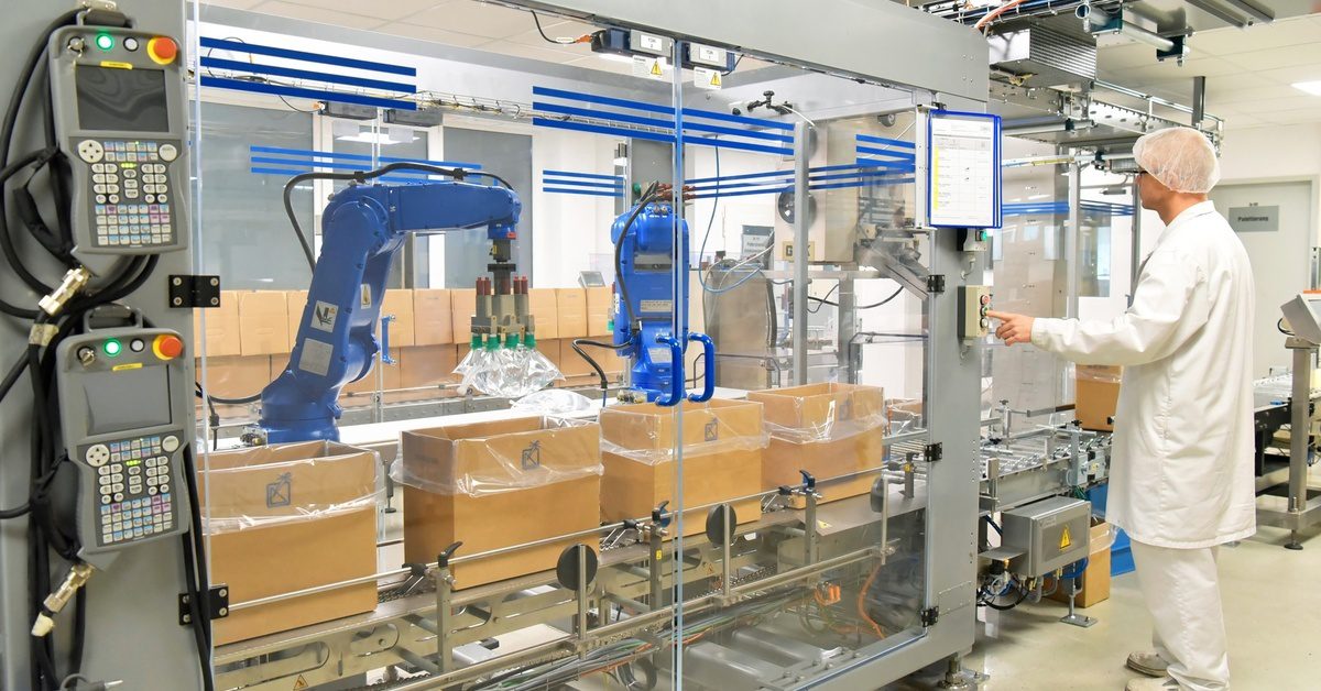 A cleanroom with blue robotic arms placing pouches into boxes on a conveyor, supervised by a worker in a lab coat.