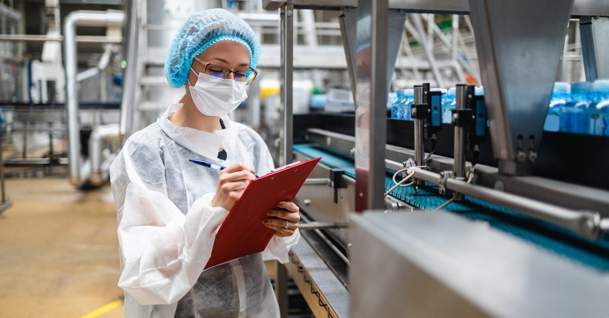 A woman in protective gear writes on a clipboard near a conveyor belt with blue bottles in a bright industrial facility.