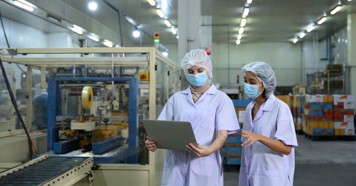 Two women wearing lab coats and masks stand in a factory, holding a tablet amidst machinery and stacked goods.