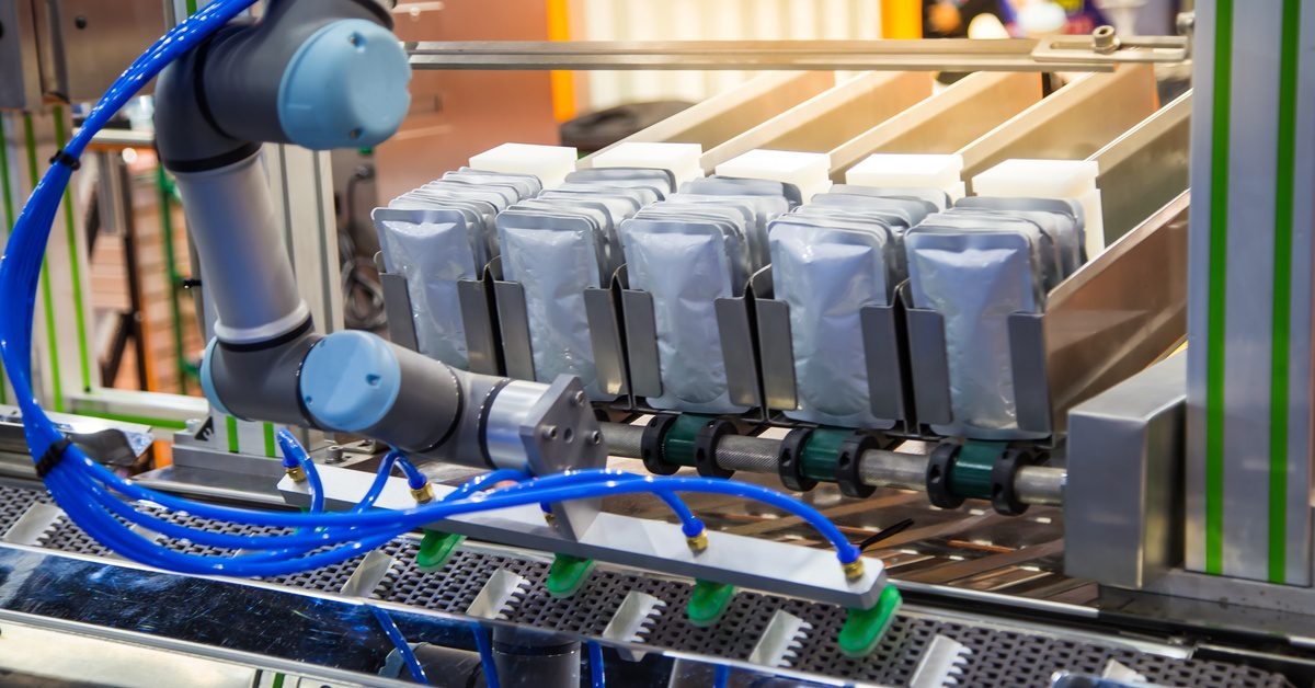 Automated packaging line with robotic arm placing sealed flexible pouches into metal holders on a conveyor system.