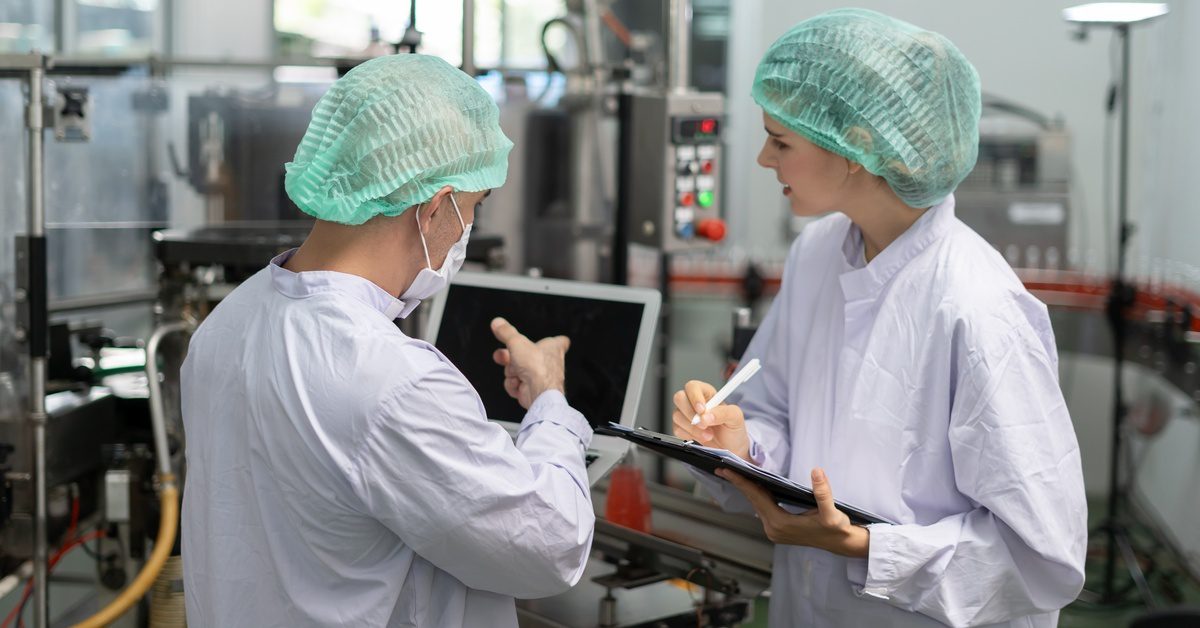 Two individuals in white coats and hairnets evaluate a machine while taking notes on a clipboard and computer.