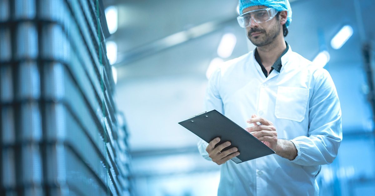Quality technician in a lab coat, hairnet, and safety glasses inspects stacked packaged trays while holding a clipboard.