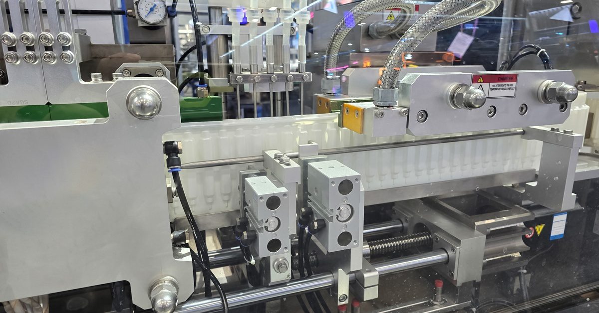 A packaging machine with pneumatic tubing, wiring, and stainless steel components visible behind a clear glass panel.