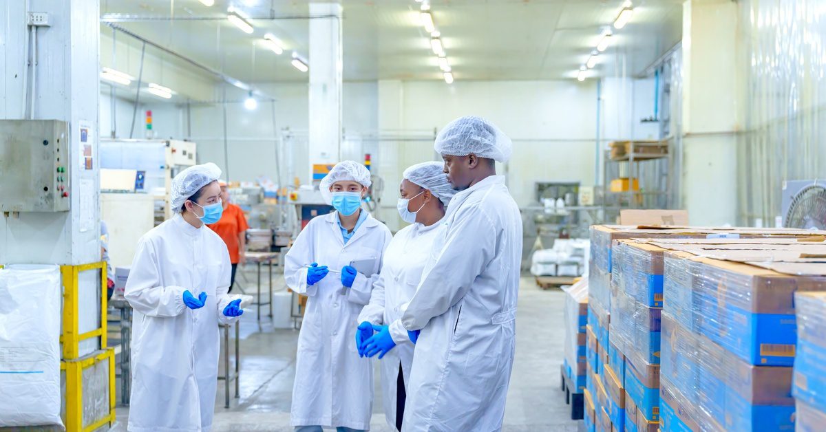 Four workers wearing white coats, face masks, and hairnets stand on a production floor next to stacks of packed boxes.