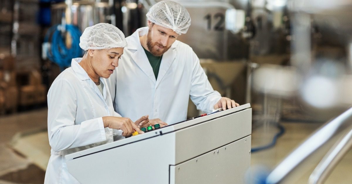 A man and a woman wearing white coats and hairnets stand in front of a control panel in an industrial plant.