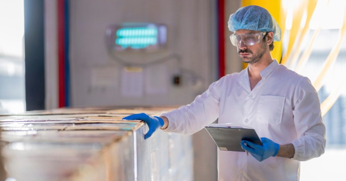 A factory worker in a white coat, blue hairnet, and gloves inspects a shipment of boxes while holding a clipboard.