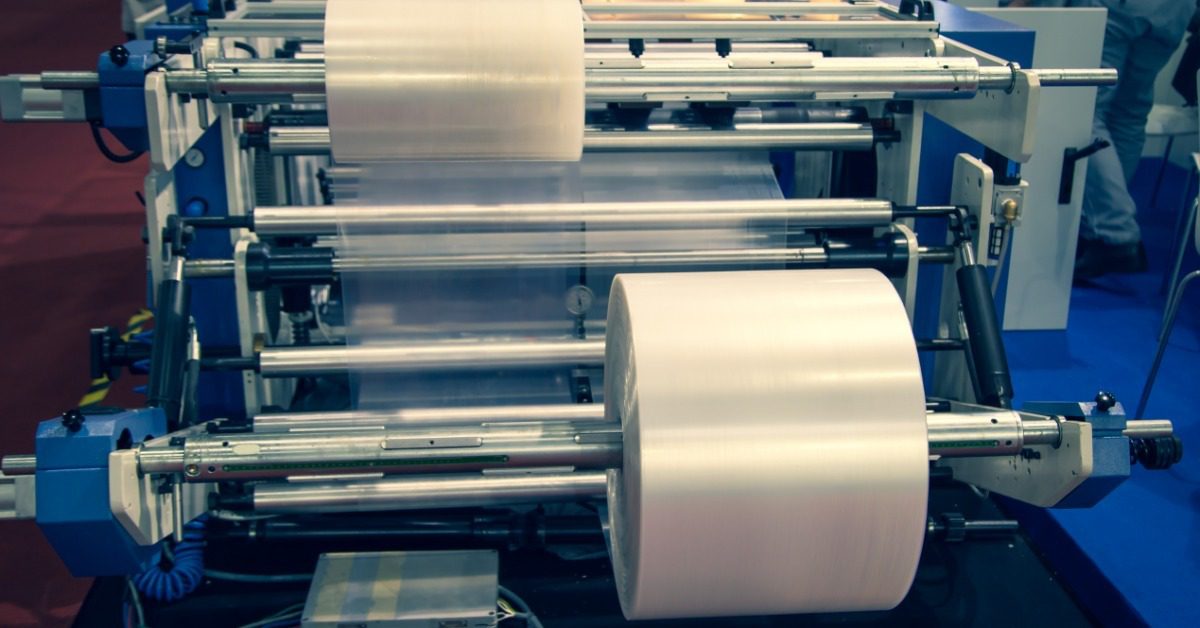 Two large rolls of transparent plastic film are being processed through a large machine in an industrial facility.