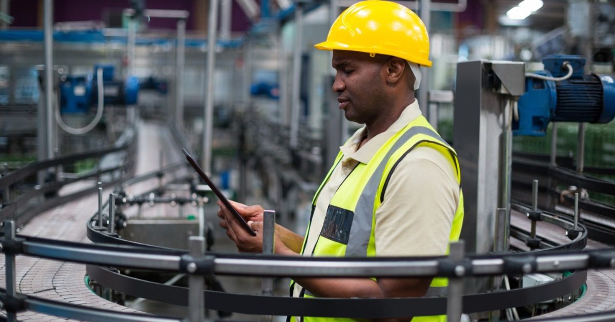 A factory worker holding a digital tablet while standing beside a production line inside an industrial facility.