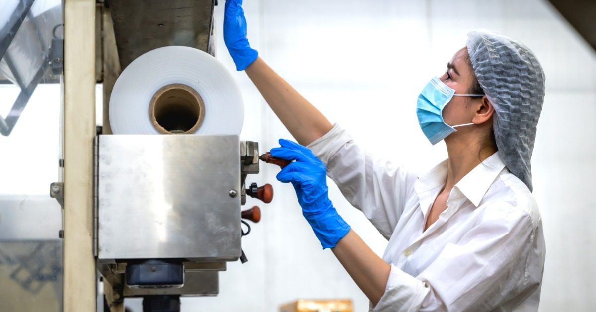 A worker handling plastic packaging materials in a manufacturing facility with equipment and containers visible.