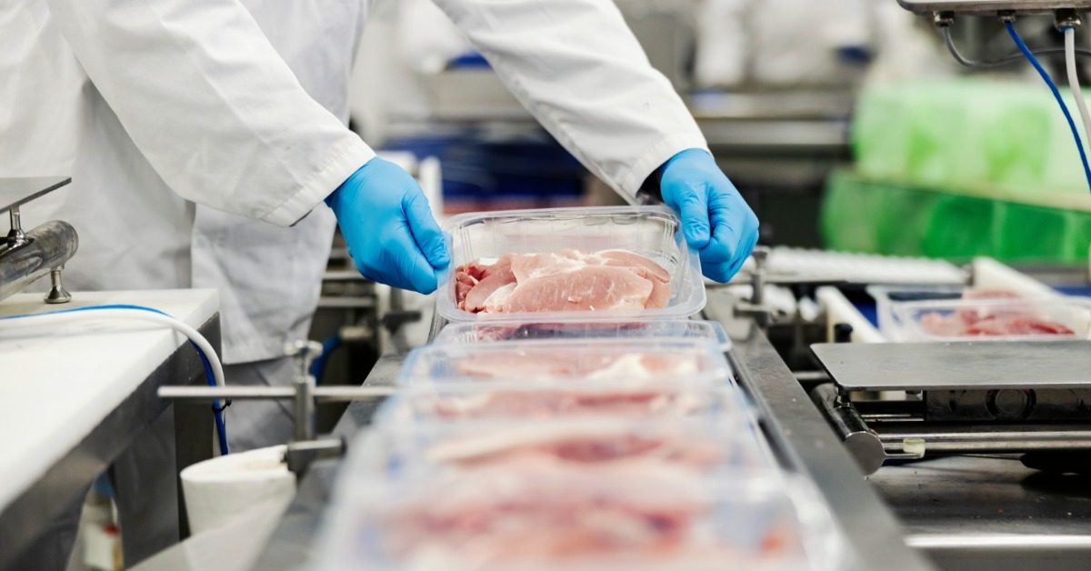 A person wearing gloves and a white lab coat gathers sealed meat packages from a conveyor belt beside other trays.